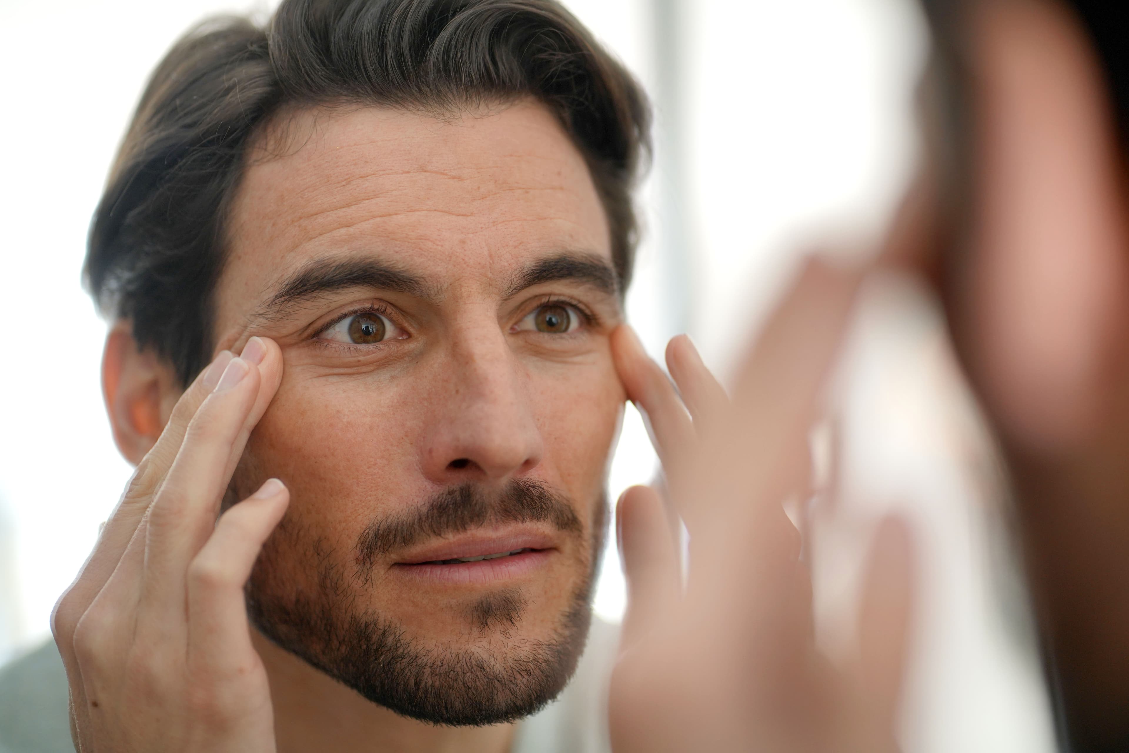 A man examining fine lines around his eyes in a mirror